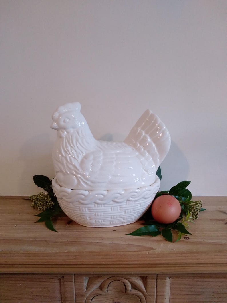 May include: A white ceramic hen-shaped egg holder with a basket-weave base. The hen is perched atop a bed of greenery, with a single brown egg nestled beside it. The piece is displayed on a wooden surface.