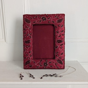 May include: A burgundy photo frame with detailed beaded floral and paisley patterns. The rectangular frame has a central aperture. A matching jewellery set, including earrings and a necklace, is displayed nearby.
