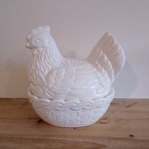 May include: A white ceramic hen-shaped container with a basket-weave base. The hen has detailed feather and facial features. The lid of the container is the hen's body, and the base is a basket. The container is sitting on a wooden surface.