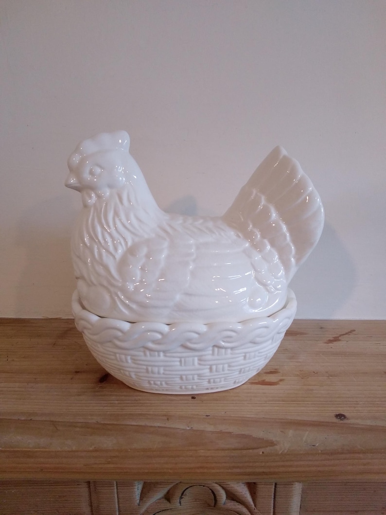 May include: A white ceramic hen-shaped container with a basket-weave base. The hen has detailed feather and facial features. The lid of the container is the hen's body, and the base is a basket. The container is sitting on a wooden surface.
