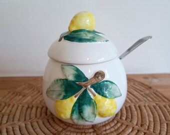 May include: White ceramic sugar bowl with a lid and a small spoon. The bowl is decorated with hand-painted lemons and green leaves. The bowl is sitting on a woven placemat.