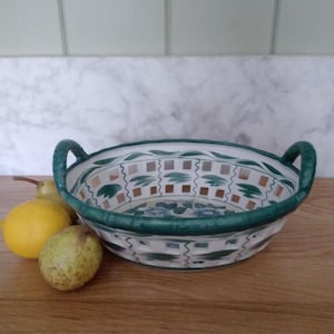 May include: A white ceramic bowl with green and brown accents. The bowl has two handles and a lattice design. It is sitting on a wooden surface with fruit.