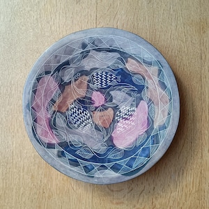May include: A round, grey ceramic plate with a white design of stylised fish and leaves. The plate has a blue and pink watercolour wash.
