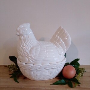 May include: A white ceramic hen-shaped egg holder with a basket-weave base. The hen sits atop the basket, with a single brown egg and greenery beside it. The holder is a decorative kitchen item.