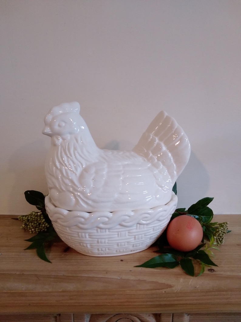 May include: A white ceramic hen-shaped egg holder with a basket-weave base. The hen sits atop the basket, with a single brown egg and greenery beside it. The holder is a decorative kitchen item.