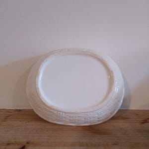 May include: A white, oval-shaped ceramic serving dish. The dish has a smooth, glossy interior and a textured, basketweave-like pattern around the rim. The dish is sitting on a wooden surface.