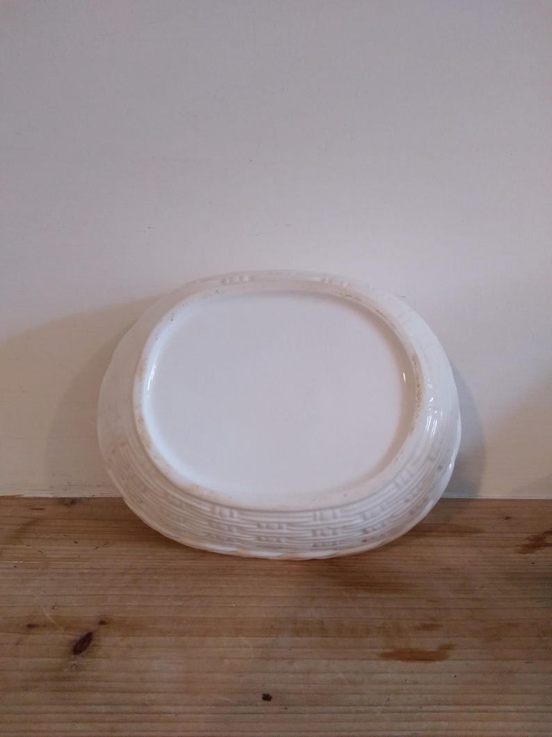May include: A white, oval-shaped ceramic serving dish. The dish has a smooth, glossy interior and a textured, basketweave-like pattern around the rim. The dish is sitting on a wooden surface.