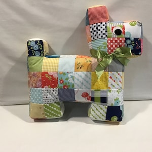 May include: A dog-shaped cushion made from patchwork fabric. The cushion features a variety of patterned squares, including floral and polka dot designs. A green ribbon bow adorns the dog's neck, and it has a black eye.