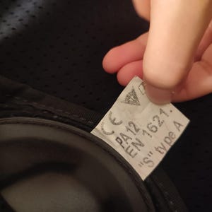 Może przedstawiać: Zbliżenie etykiety z napisem "CE PA12 EN 1621. 'S' type A" na czarnej powierzchni. Etykieta jest trzymana przez dłoń. Etykieta ma białe tło i czarny tekst.