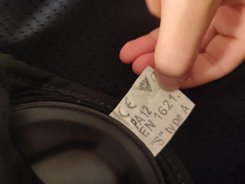 Może przedstawiać: Zbliżenie etykiety z napisem "CE PA12 EN 1621. 'S' type A" na czarnej powierzchni. Etykieta jest trzymana przez dłoń. Etykieta ma białe tło i czarny tekst.