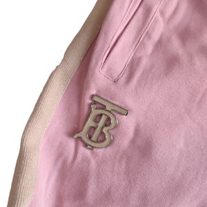 Puede incluir: Primer plano de un pantal&oacute;n de ch&aacute;ndal rosa con un logotipo plateado. El pantal&oacute;n de ch&aacute;ndal tiene un bolsillo y una franja beige en el lateral. El logotipo es una letra "B" estilizada.
