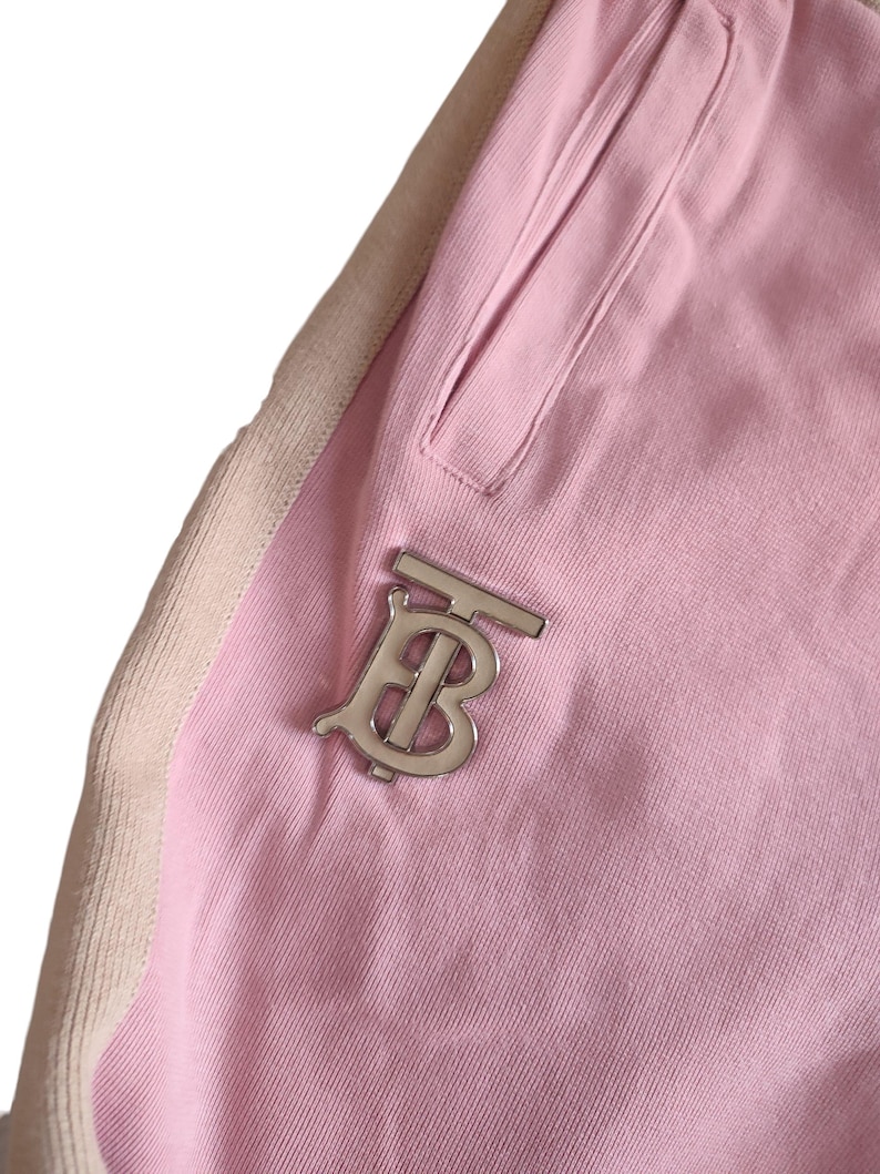 Puede incluir: Primer plano de un pantal&oacute;n de ch&aacute;ndal rosa con un logotipo plateado. El pantal&oacute;n de ch&aacute;ndal tiene un bolsillo y una franja beige en el lateral. El logotipo es una letra "B" estilizada.