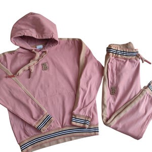 Puede incluir: Conjunto de ch&aacute;ndal rosa compuesto por una sudadera con capucha y un pantal&oacute;n de ch&aacute;ndal. La sudadera tiene ribetes beige, una capucha con cord&oacute;n y un logotipo. El pantal&oacute;n de ch&aacute;ndal tiene cintura con cord&oacute;n y ribetes a juego.