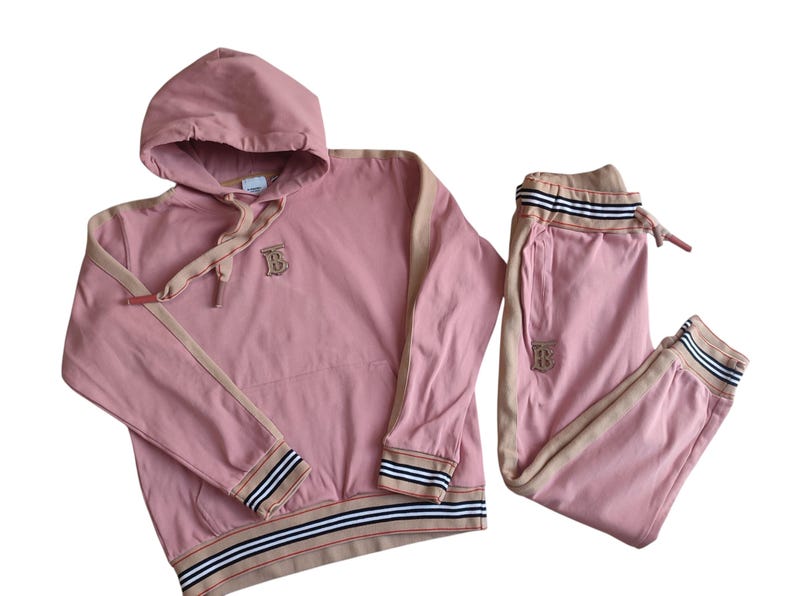 Puede incluir: Conjunto de ch&aacute;ndal rosa compuesto por una sudadera con capucha y un pantal&oacute;n de ch&aacute;ndal. La sudadera tiene ribetes beige, una capucha con cord&oacute;n y un logotipo. El pantal&oacute;n de ch&aacute;ndal tiene cintura con cord&oacute;n y ribetes a juego.