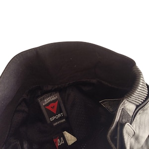 Może przedstawiać: Czarna sk&oacute;rzana kurtka motocyklowa z kołnierzem i zamkiem błyskawicznym. Kurtka posiada metkę "DAINESE SAFETY EQUIPMENT" ze słowem "SPORT" oraz metkę z rozmiarem 44. Wyprodukowano na Ukrainie.