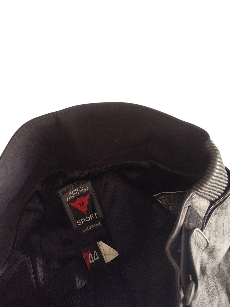 Może przedstawiać: Czarna sk&oacute;rzana kurtka motocyklowa z kołnierzem i zamkiem błyskawicznym. Kurtka posiada metkę "DAINESE SAFETY EQUIPMENT" ze słowem "SPORT" oraz metkę z rozmiarem 44. Wyprodukowano na Ukrainie.