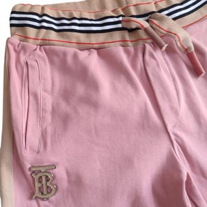 Puede incluir: Pantalones deportivos rosas con una raya beige en el lateral y una cintura beige con rayas blancas y negras. Los pantalones deportivos tienen un logotipo plateado y un cord&oacute;n. El material parece suave y c&oacute;modo.