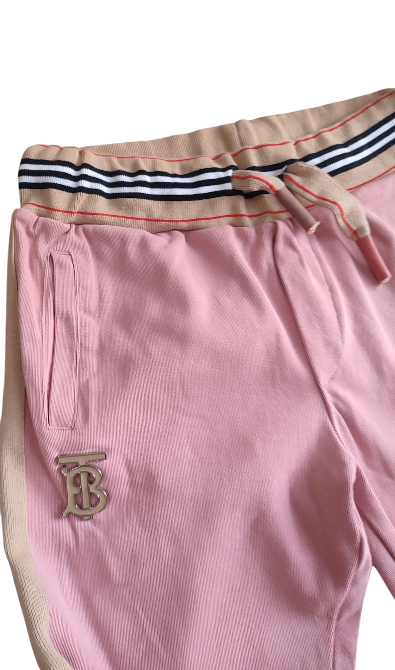 Puede incluir: Pantalones deportivos rosas con una raya beige en el lateral y una cintura beige con rayas blancas y negras. Los pantalones deportivos tienen un logotipo plateado y un cord&oacute;n. El material parece suave y c&oacute;modo.