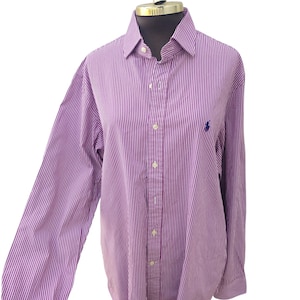 Può includere: Camicia button-down a righe viola e bianche con maniche lunghe. La camicia presenta un piccolo logo blu sul petto. La camicia è esposta su un manichino.