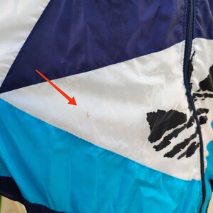 May include: A close-up of a zipped jacket with a colour block design. The jacket features sections of white, navy blue, and turquoise. Black abstract shapes are also visible on the jacket.