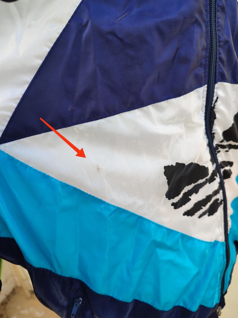 May include: A close-up of a zipped jacket with a colour block design. The jacket features sections of white, navy blue, and turquoise. Black abstract shapes are also visible on the jacket.