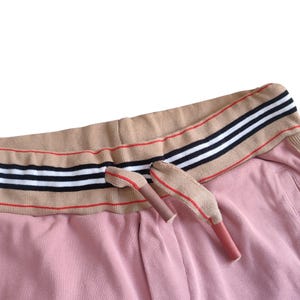 Puede incluir: Primer plano de un pantal&oacute;n de ch&aacute;ndal rosa con una cintura color canela con rayas negras, blancas y rojas. Los extremos del cord&oacute;n son color canela con detalles rojos. La tela tiene una textura acanalada.