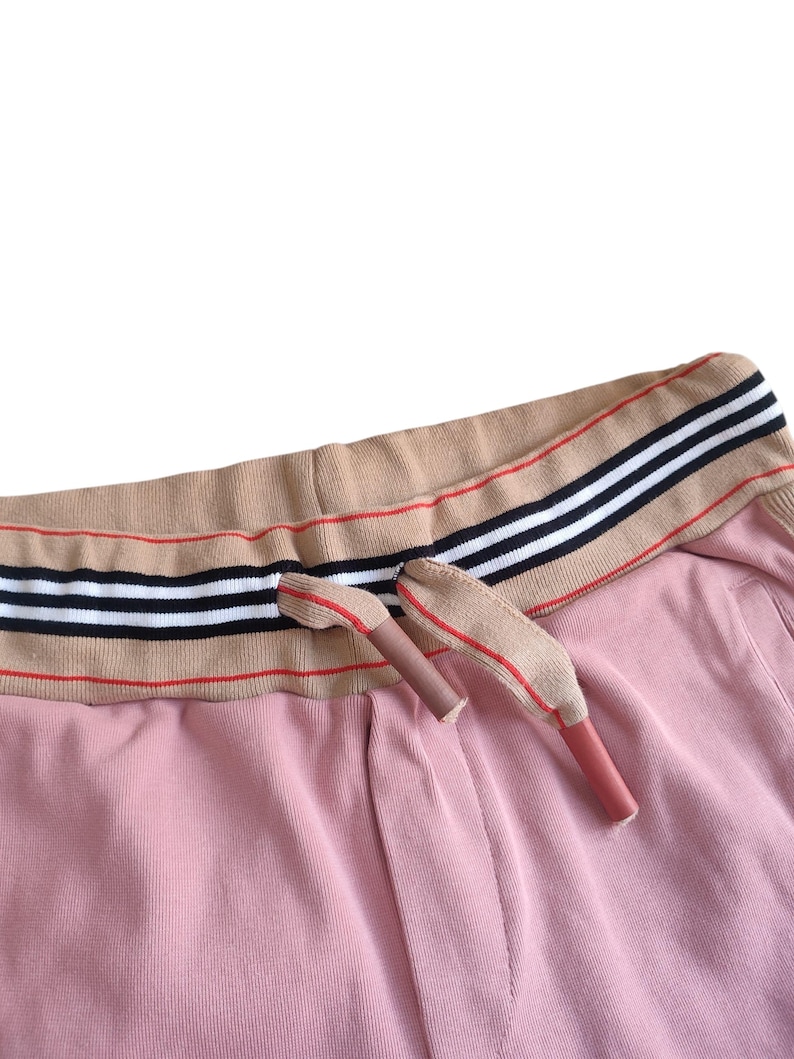 Puede incluir: Primer plano de un pantal&oacute;n de ch&aacute;ndal rosa con una cintura color canela con rayas negras, blancas y rojas. Los extremos del cord&oacute;n son color canela con detalles rojos. La tela tiene una textura acanalada.