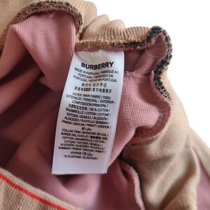 Puede incluir: Primer plano de una prenda beige y rosa con una etiqueta. La etiqueta dice "BURBERRY" y enumera la composici&oacute;n de la tela. La prenda tiene un ribete a rayas con rayas rojas, negras y blancas.