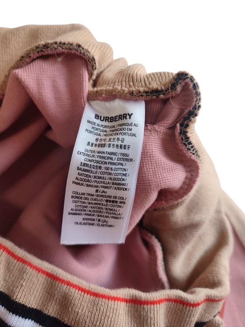 Puede incluir: Primer plano de una prenda beige y rosa con una etiqueta. La etiqueta dice "BURBERRY" y enumera la composici&oacute;n de la tela. La prenda tiene un ribete a rayas con rayas rojas, negras y blancas.
