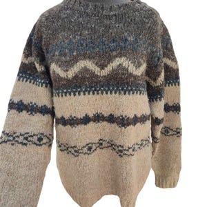 May include: A brown and beige sweater with a patterned design. The sweater features a crew neck and long sleeves. The design includes blue, brown, and beige zigzag and diamond patterns across the chest and sleeves. The sweater is made of a thick, textured material.