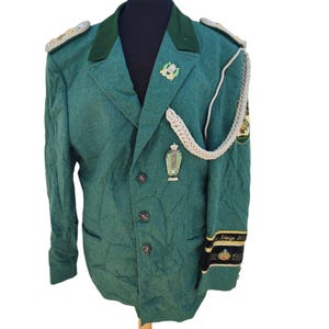 May include: A green vintage jacket with a dark green collar and lapels, displayed on a mannequin. The jacket features decorative shoulder boards, a silver cord, and multiple emblems. The sleeve has text and the years 1982 and 1991.