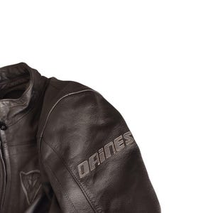 Może przedstawiać: Zbliżenie czarnej sk&oacute;rzanej kurtki motocyklowej. Kurtka posiada nazwę marki "DAINESE" w srebrnych literach na rękawie. Srebrne logo jest widoczne na piersi. Kurtka ma wysoki kołnierz i widoczne szwy.