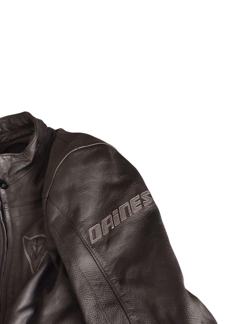 Może przedstawiać: Zbliżenie czarnej sk&oacute;rzanej kurtki motocyklowej. Kurtka posiada nazwę marki "DAINESE" w srebrnych literach na rękawie. Srebrne logo jest widoczne na piersi. Kurtka ma wysoki kołnierz i widoczne szwy.