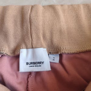 Puede incluir: Primer plano de un pantal&oacute;n de ch&aacute;ndal rosa y beige con una etiqueta blanca que dice "BURBERRY LONDON ENGLAND". El pantal&oacute;n tiene una cintura el&aacute;stica y una tela suave y texturizada.