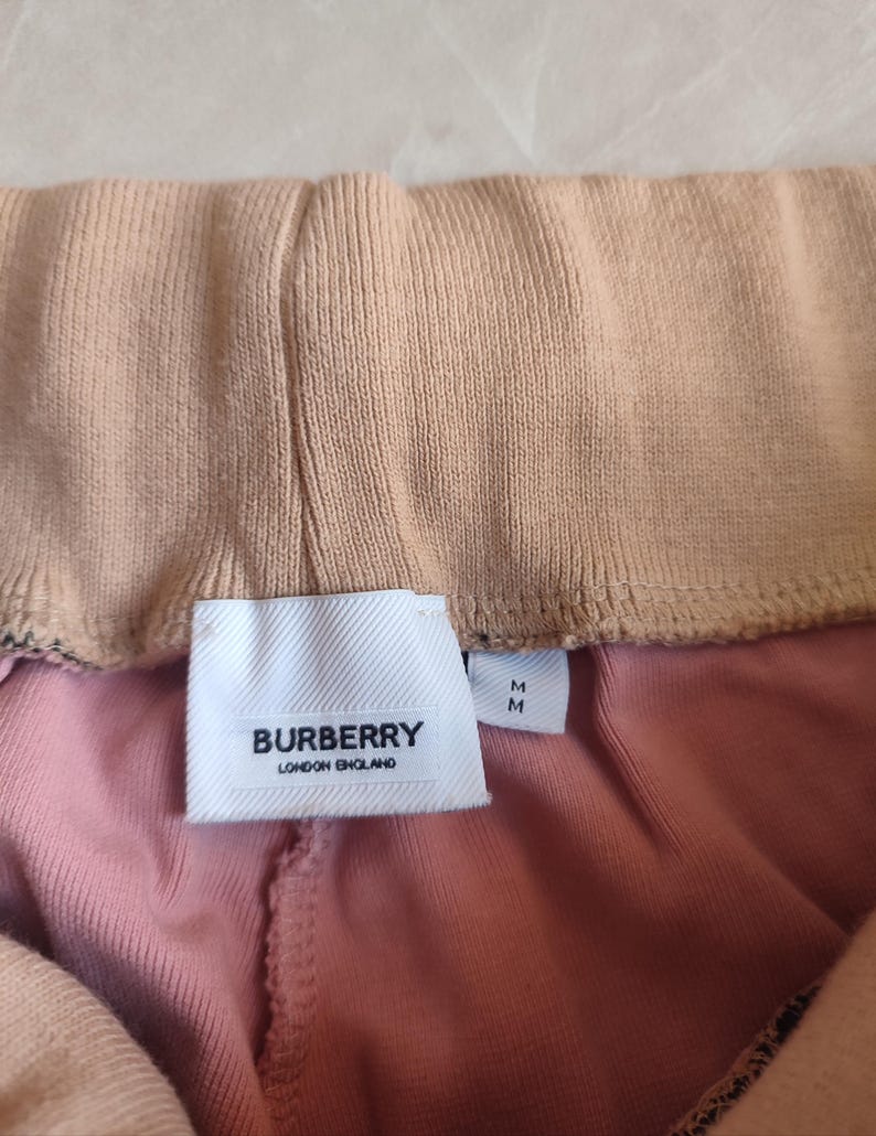 Puede incluir: Primer plano de un pantal&oacute;n de ch&aacute;ndal rosa y beige con una etiqueta blanca que dice "BURBERRY LONDON ENGLAND". El pantal&oacute;n tiene una cintura el&aacute;stica y una tela suave y texturizada.