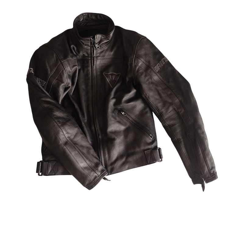 Może przedstawiać: Czarna sk&oacute;rzana kurtka motocyklowa z wysokim kołnierzem, zapięciem na zamek błyskawiczny i wieloma kieszeniami. Kurtka ma nazwę marki "DAINESE" na rękawach i logo na piersi. Posiada regulowane paski w talii.