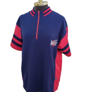 Peut inclure: Un t-shirt bleu marine et rouge à manches courtes avec un col zippé. Le t-shirt présente des manches rouges avec des rayures bleu marine et un logo Rockets sur la poitrine. Le t-shirt est présenté sur un mannequin.