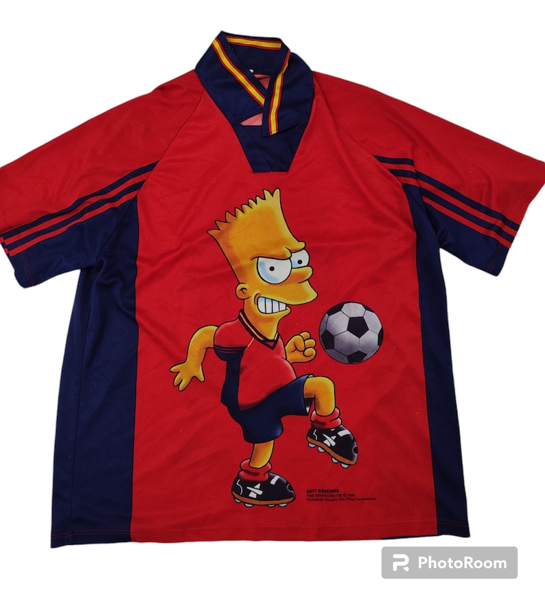 May include: Red and navy blue soccer jersey featuring Bart Simpson kicking a soccer ball. The jersey has a V-neck collar with yellow and red trim, and navy blue stripes on the sleeves. The jersey has a cartoon graphic of Bart Simpson.
