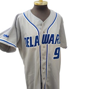 Peut inclure: Un maillot de baseball gris avec le mot "DELAWARE" en lettres bleues et blanches sur la poitrine. Le numéro "9" est sur le devant. Des bordures bleues et blanches ornent le col et les manches. Le maillot est boutonné.