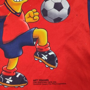 May include: Red jersey featuring a cartoon character kicking a soccer ball. The character is wearing a red and blue uniform. The jersey has the text "Matt Groening The Simpsons TM 1998 Twentieth Century Fox Film Corporation."