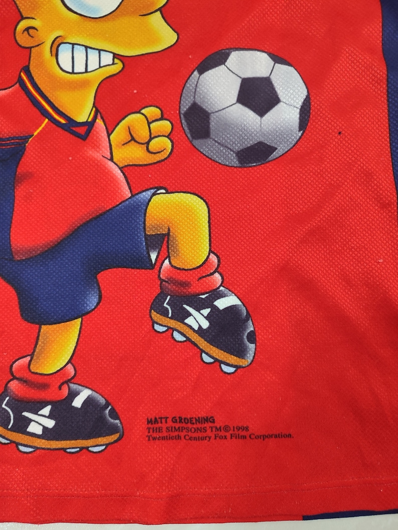 May include: Red jersey featuring a cartoon character kicking a soccer ball. The character is wearing a red and blue uniform. The jersey has the text "Matt Groening The Simpsons TM 1998 Twentieth Century Fox Film Corporation."