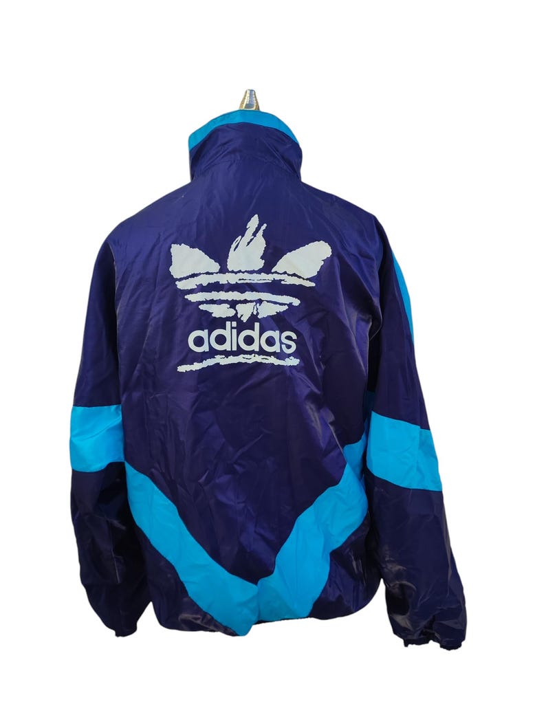 May include: A vintage Adidas track jacket in a striking colour scheme. The jacket has a deep purple base with turquoise blue accents on the sleeves and sides. The Adidas logo is displayed in white on the back.