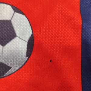 May include: Close-up of a red athletic jersey with a soccer ball graphic. The jersey has a mesh texture and a blue stripe on the side. A small black dot is visible on the red fabric.