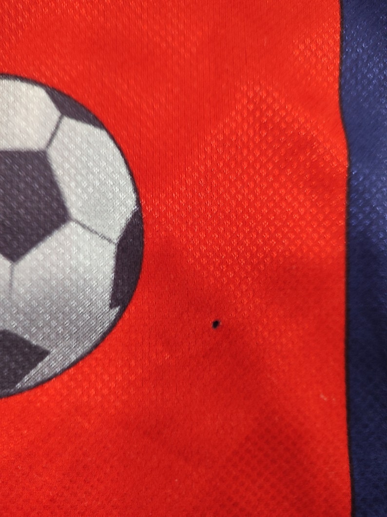 May include: Close-up of a red athletic jersey with a soccer ball graphic. The jersey has a mesh texture and a blue stripe on the side. A small black dot is visible on the red fabric.