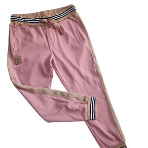 Puede incluir: Pantalones deportivos rosas con cintura con cord&oacute;n y bolsillos laterales. Los pantalones tienen una franja beige en los laterales y alrededor de la cintura y los tobillos. Un logotipo bordado dorado es visible en la pierna izquierda. La cintura tiene un dise&ntilde;o a rayas.