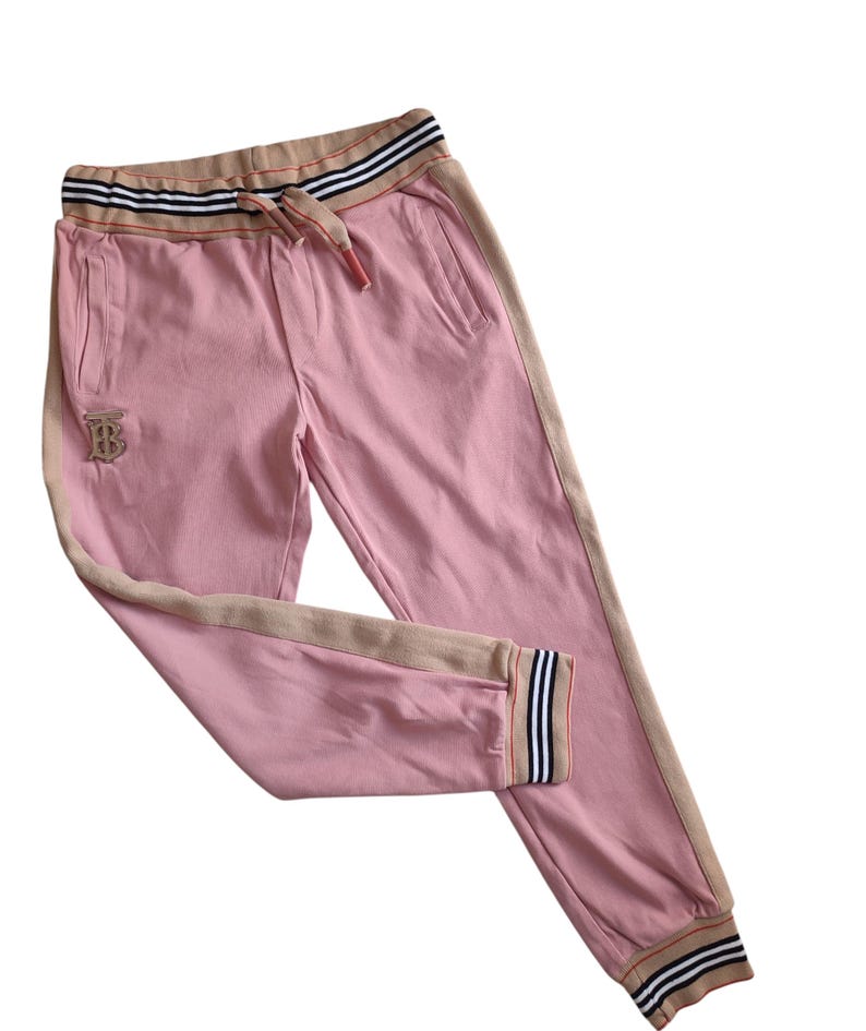 Puede incluir: Pantalones deportivos rosas con cintura con cord&oacute;n y bolsillos laterales. Los pantalones tienen una franja beige en los laterales y alrededor de la cintura y los tobillos. Un logotipo bordado dorado es visible en la pierna izquierda. La cintura tiene un dise&ntilde;o a rayas.