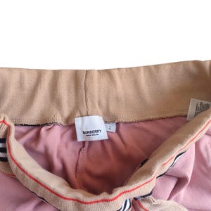 Puede incluir: Primer plano de un pantal&oacute;n de ch&aacute;ndal rosa con cord&oacute;n y cintura beiges. El cord&oacute;n tiene rayas rojas y negras. Se ve una etiqueta blanca con la marca "BURBERRY LONDON ENGLAND".