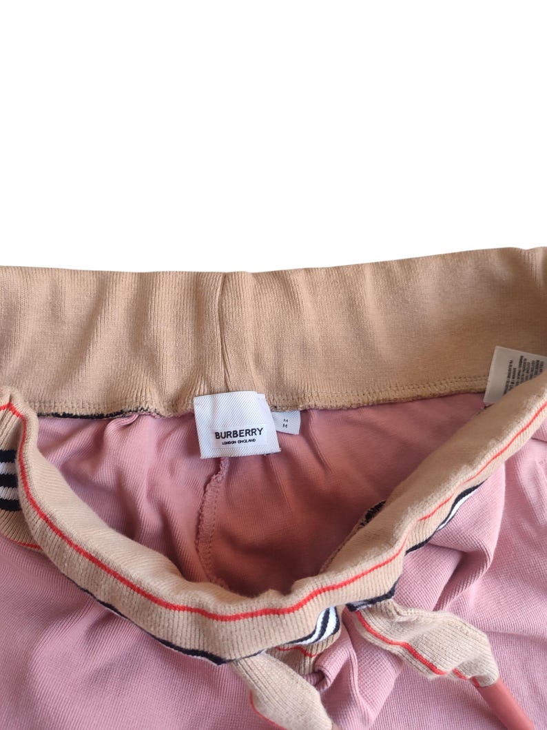 Puede incluir: Primer plano de un pantal&oacute;n de ch&aacute;ndal rosa con cord&oacute;n y cintura beiges. El cord&oacute;n tiene rayas rojas y negras. Se ve una etiqueta blanca con la marca "BURBERRY LONDON ENGLAND".