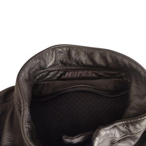 Może przedstawiać: Zbliżenie czarnego sk&oacute;rzanego kołnierza kurtki motocyklowej. Kurtka posiada zamek błyskawiczny z przodu i podszewkę z siatki. Nazwa marki "DAINESE" jest widoczna na kołnierzu. Sk&oacute;ra ma teksturowany wygląd.