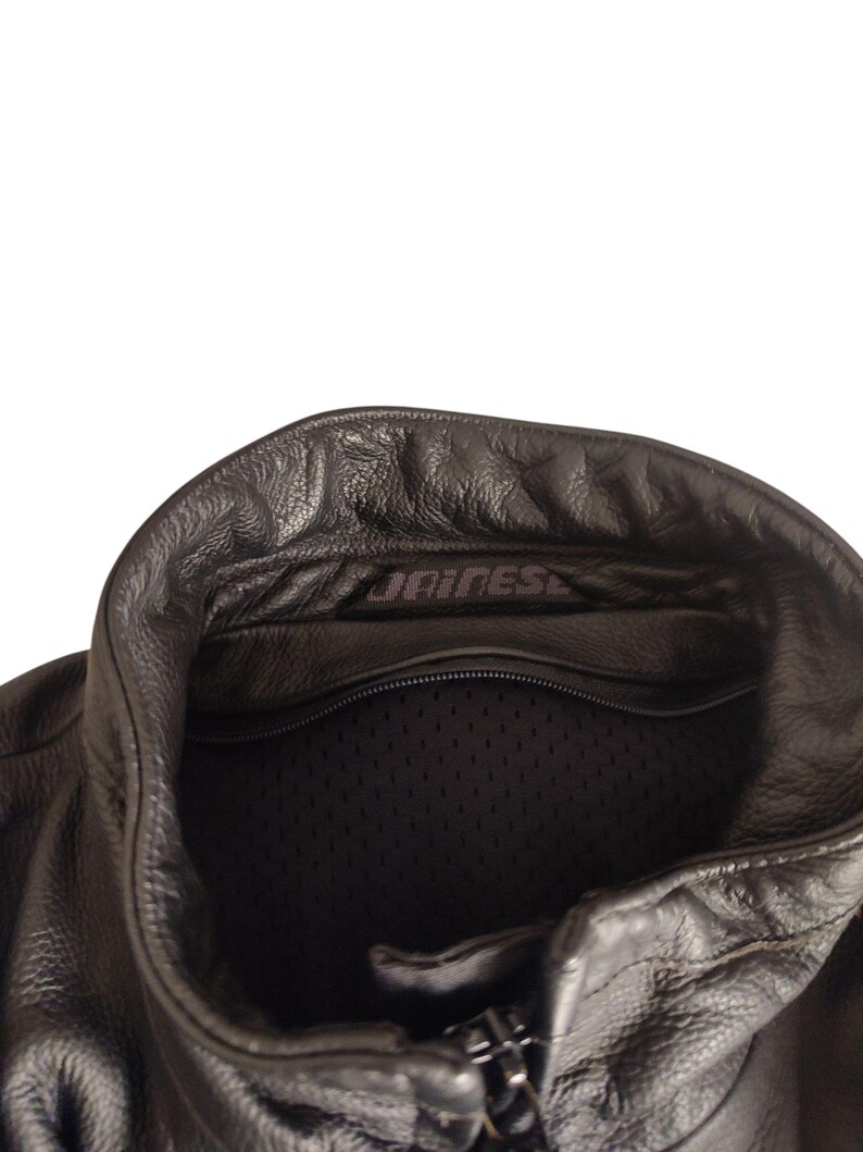 Może przedstawiać: Zbliżenie czarnego sk&oacute;rzanego kołnierza kurtki motocyklowej. Kurtka posiada zamek błyskawiczny z przodu i podszewkę z siatki. Nazwa marki "DAINESE" jest widoczna na kołnierzu. Sk&oacute;ra ma teksturowany wygląd.